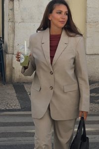 Μπεζ σακάκι με δύο κουμπιά και εμπρός τσέπες, σε συνδυασμό με το αντίστοιχο παντελόνι. Φοριέται πάνω από ένα κόκκινο τοπ, κρατώντας ένα πράσινο ποτό και μια μαύρη τσάντα.