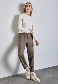 Sweatshirt beige clair avec texte en relief ; pantalons vert olive avec poches latérales et accessoires en argent ; chaussures de sport beige et blanche.