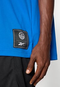 Zila sporta krekla ar teksturētu audumu malu ir melna taisnstūra uzlīme, kurā attēlots balts basketbola grafiskais attēls un logo.