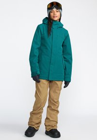 Veste imperméable bleu-vert avec capuche et poches latérales, associée à un pantalon de neige beige. Des gants noirs et des lunettes de ski complètent la tenue d'hiver.