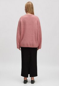Overmaatse roze gebreide trui met verlaagde schouders, gecombineerd met een zwarte lange rok met een zijsplit. Het model draagt zwarte schoenen.