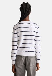 Biały sweter w granatowe poziome paski, z prążkowanymi mankietami i dołem, o teksturowanym wzorze, noszony z szarymi spodniami, widok z tyłu.