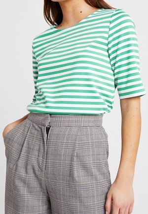 Žena v zeleno-bílém tričku s vodorovným proužkem a vysokým pasem šedých kostkovaných kalhotách s předním zipem a kapsami.