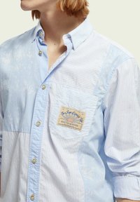 Camicia azzurro chiaro con strisce e tasca a toppa con etichetta. Colletto button-down, texture morbida e dettagli di cucitura visibili.