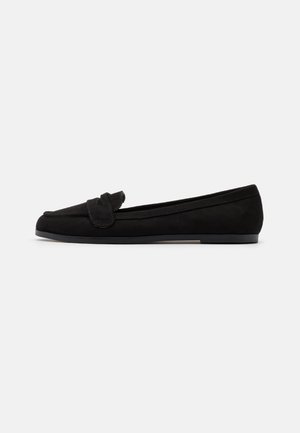 Mocasín plano de ante negro con tacón bajo y detalle de tira a juego en la parte frontal, mostrado de perfil sobre un fondo liso.