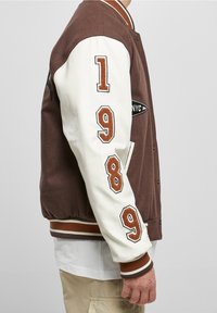 Brun och vit varsityjacka med broderade siffrorna "1989" på ärmarna, svart logotyp-patch på bröstet, ribbade mudd och nederkant.