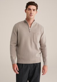 Hellgrauer Zip-Neck-Pullover mit strukturierter Oberfläche, ausgestattet mit einer kleinen Brusttasche und gerippten Bündchen, kombiniert mit dunklen Hosen.