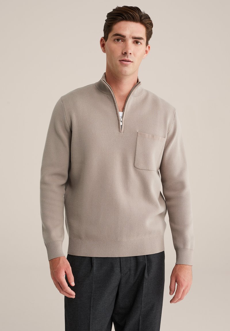 Hellgrauer Zip-Neck-Pullover mit strukturierter Oberfläche, ausgestattet mit einer kleinen Brusttasche und gerippten Bündchen, kombiniert mit dunklen Hosen.