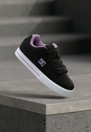 Baskets noires avec doublure et logo lavande, semelle blanche avec une bande lavande, flottant au-dessus d'une marche en pierre dans un environnement extérieur.