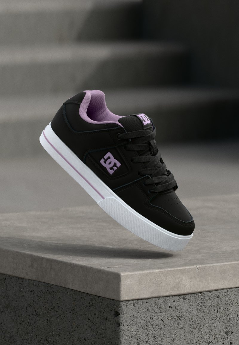 Μαύρο αθλητικό παπούτσι με μωβ επένδυση και λογότυπο, λευκή σόλα με μωβ ρίγα, αιωρούμενο πάνω από λιθόστρωτη σκάλα σε εξωτερικό χώρο.