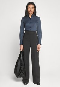 Camicia in satin navy con bottoni abbinata a pantaloni larghi neri a vita alta. Il modello tiene una borsa in pelle nera. Design semplice e strutturato.