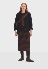 Donkerblauwe jasje met knopen, bordeaux top, geruite maxi-rok in rood en donkerblauw, en kniehoge bruine laarzen. Heeft een crossbodytas.