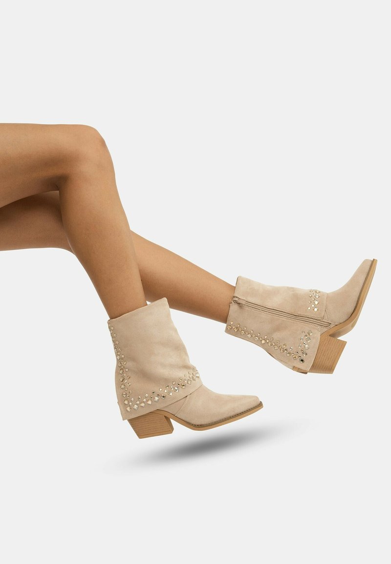 Botas de tobillo de ante beige con tacón cuadrado, que presentan tachuelas decorativas a lo largo del costado y un borde superior con forma de ondas.