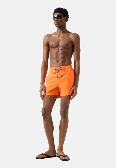 Jonge man draagt feloranje zwemshorts, zwarte slippers en zwarte zonnebril, staand tegen een effen lichte achtergrond.