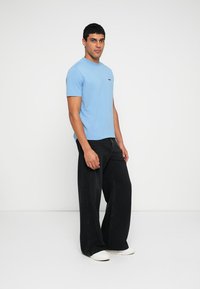 Mėlyna medvilninė trumpomis rankovėmis marškinėliai su mažu logotipu, derinami su juodais plačiomis kelnėmis džinsais ir baltais sportbačiais ant vienspalvio fono.