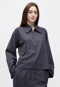 Mladá žena s dlhými hnedými vlasmi, oblečená v voľnej tmavosivej košeli s golierom a zladených nohaviciach, ruky vreckách, neutrálne pozadie.