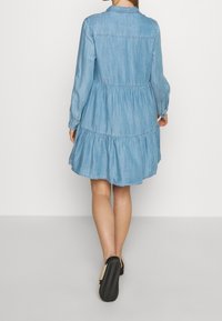 Vaaleansininen farkkumekko, jossa on pitkät hihat ja kerroksellinen, väljät hameosa. Mekossa on kaulus ja nappisuljenta kaulassa. Litteät mustat kengät.