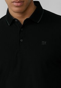Schwarzes Poloshirt aus strukturiertem Stoff. Verfügt über eine Dreiknopf-Leiste, ein dezentes Logo und kontrastierende Kragenverzierungen.
