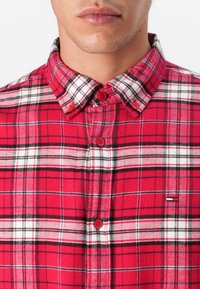 Camisa de franela de cuadros rojos con líneas negras y blancas, diseño de botones en el frente y un pequeño logo en el lado izquierdo del pecho. Textura suave.