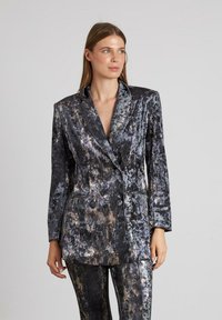 Chaqueta de corte cruzado en tejido texturizado negro y gris con patrones abstractos, que presenta un collar afilado y bolsillos laterales.