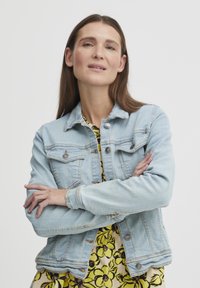 Šviesiai mėlynas džinsinis švarkas su mygtukais priekyje, krūtinės kišenėmis ir prigludusia forma, dėvimas virš gėlių rašto geltonos suknelės.