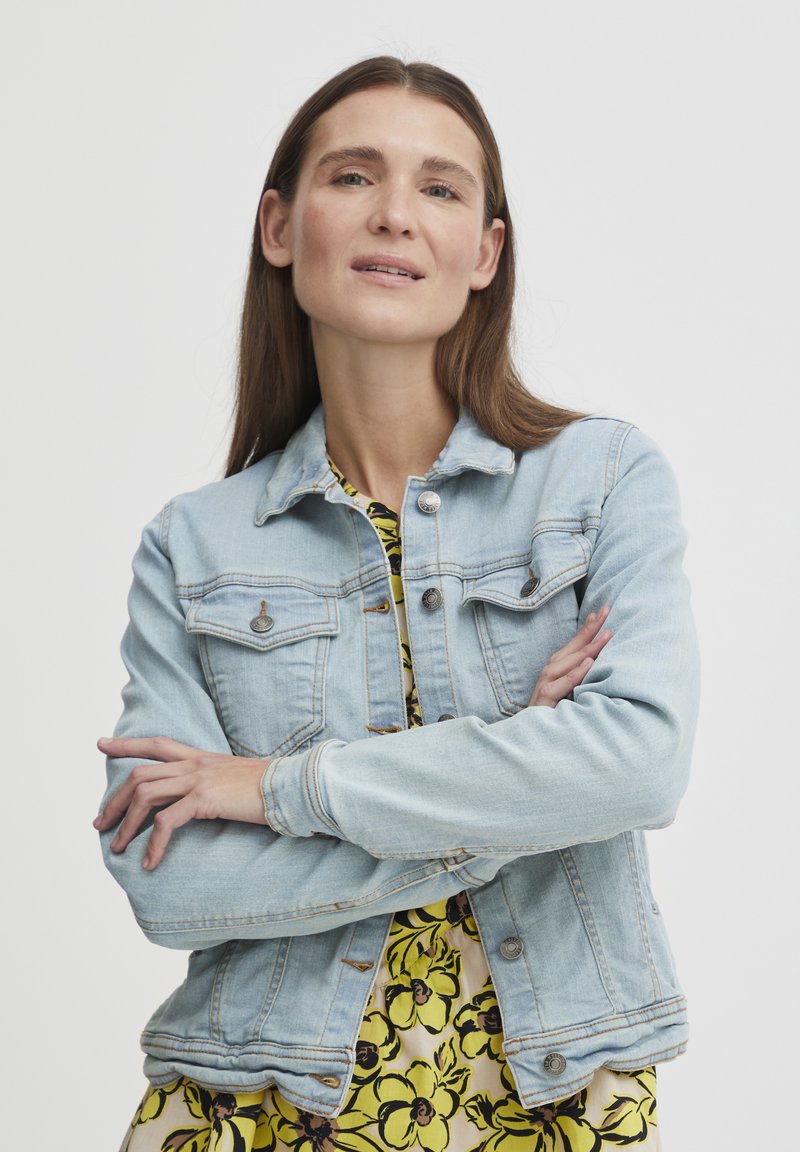 Šviesiai mėlynas džinsinis švarkas su mygtukais priekyje, krūtinės kišenėmis ir prigludusia forma, dėvimas virš gėlių rašto geltonos suknelės.