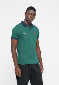 Zaļa polo krekls ar tumši zilu apkakli un piedurkņu akcentiem. Uz krūšu ir neliels logotips. Audums izskatās gluds un viegls.