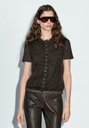 Brown fuzzy knit vest with round neckline and button-down front, paired with a black T-shirt and dark leather pants. Model wears sunglasses.