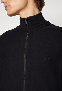 Svart zip-up tröja med hög ribbad krage, med en diskret logotyp på bröstet och slät stickad textur. Dragkedjan går centralt.