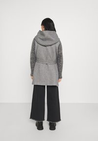 Cappotto grigio con un ampio cappuccio, maniche grigie testurizzate e una cintura, abbinato a pantaloni neri a gamba larga e stivali neri.