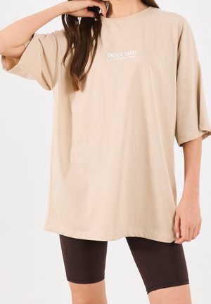 Vrouw draagt een oversized beige T-shirt met de tekst "Pacific Coast California" en zwarte biker shorts op een witte achtergrond.