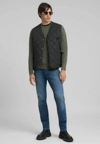 Gilet nero trapuntato sopra un maglione verde a trecce, abbinato a jeans blu e stivaletti neri. Completo con occhiali da sole; abbigliamento casual.