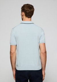 Camisa polo azul claro de mangas cortas, con cuello y un ribete negro en contraste a lo largo del cuello. El material parece suave y tiene un diseño ajustado.