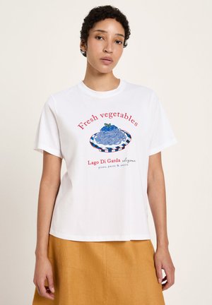 Young person wearing white t-shirt with blue pasta plate illustration and red text, paired with mustard-yellow skirt, standing against plain background.