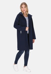 Cappotto lungo blu navy con zip davanti, cappuccio regolabile e tasche laterali. Indossato sopra una camicia a righe e abbinato a jeans e sneakers bianche.
