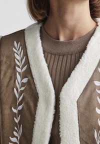 Brunt väst med vit konstpälskrage, med broderad bladdesign och ribbad brun tröja under. Mjuk textur, varmt utseende.