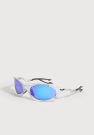 Sportowe okulary przeciwsłoneczne typu wraparound z refleksyjnymi niebieskimi soczewkami i przezroczystą oprawką z czarnymi zakrzywionymi zausznikami, spoczywające na białej powierzchni.