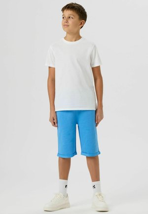 Garçon portant un t-shirt blanc uni, un short bleu jusqu'aux genoux, des chaussettes blanches et des baskets blanches, regardant vers sa gauche.