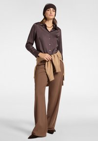 Camicia button-up in satin marrone, sovrapposta a un maglione beige annodato in vita, abbinata a pantaloni a gamba larga marroni e scarpe a punta marroni.