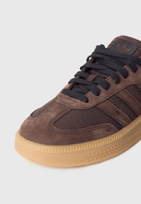 Brązowy zamszowy sneaker z czarnymi panelami z siateczki, czarnymi sznurowadłami oraz podeszwą z gumy o strukturalnym wzorze. Detale obejmują trzy paski i logo.