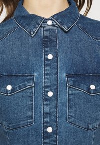 Jeansskjorta med krage i blått, med vita knappar och två bröstfickor. Synliga sömmar framhäver designen.