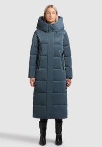 Pitkä, toppatakki tummanvihreänä, jossa korkea kaulus ja suuri huppu. Takissa on tikatut osiot ja sivutaskut. Yhdistetty mustiin saappaisiin.