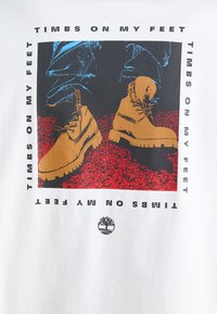 Hvit t-skjorte med en illustrasjon av brune støvler med svarte såler, blå jeans, og teksten "TIMBS ON MY FEET" i svart rundt bildet.