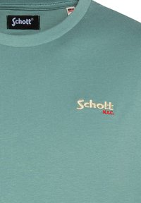 Camiseta verde de cuello redondo con un pequeño logotipo bordado "Schott N.Y.C." en beige y rojo en el pecho cerca del cuello.