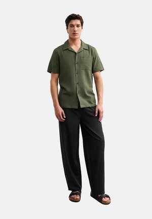 Chemise à manches courtes de couleur olive avec un devant à boutons et une poche, associée à un pantalon noir ample et des sandales sombres. Design simple.