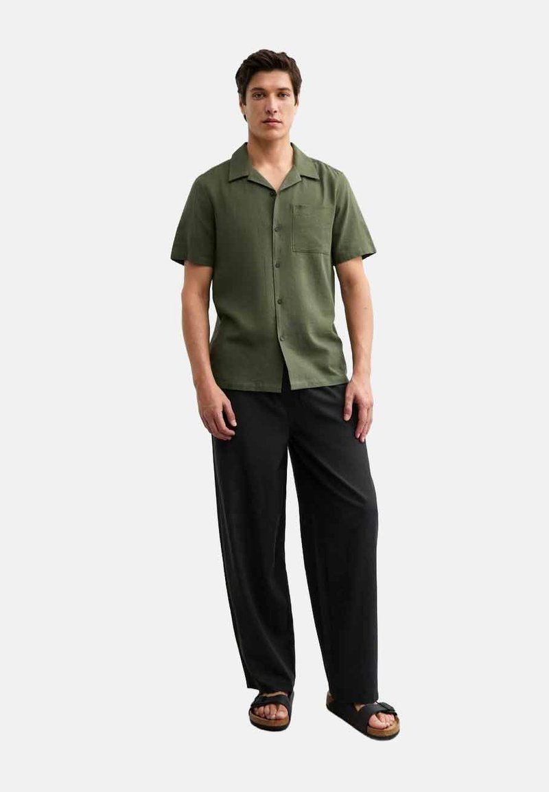 Chemise à manches courtes de couleur olive avec un devant à boutons et une poche, associée à un pantalon noir ample et des sandales sombres. Design simple.