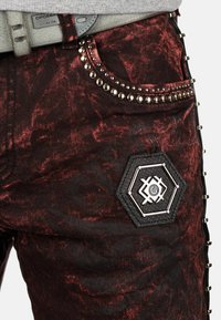 Jean en denim noir et rouge à motifs avec des clous en argent, un patch hexagonal et des détails texturés sur le côté. Ceintures incluses.