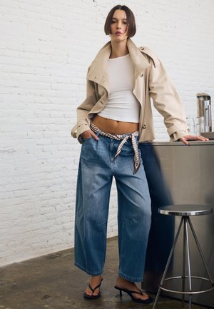 Žena v béžovom krátkom saku, bielom tielku, širokých džínsoch s vzorovaným opaskom a čiernych sandáloch na opätku sa opiera o kovový pult.