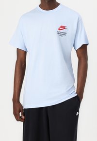 Muž v světle modrém tričku Nike s červeným logem a textem „Pouze pro autorizovaný personál“, v kombinaci s černými šortkami Nike.