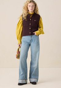 Donker bordeaux kleurige mouwloze vest met twee borstzakken en gouden knopen, gedragen over een felgele blouse met een gerimpelde kraag. Wijde blauwe jeans.